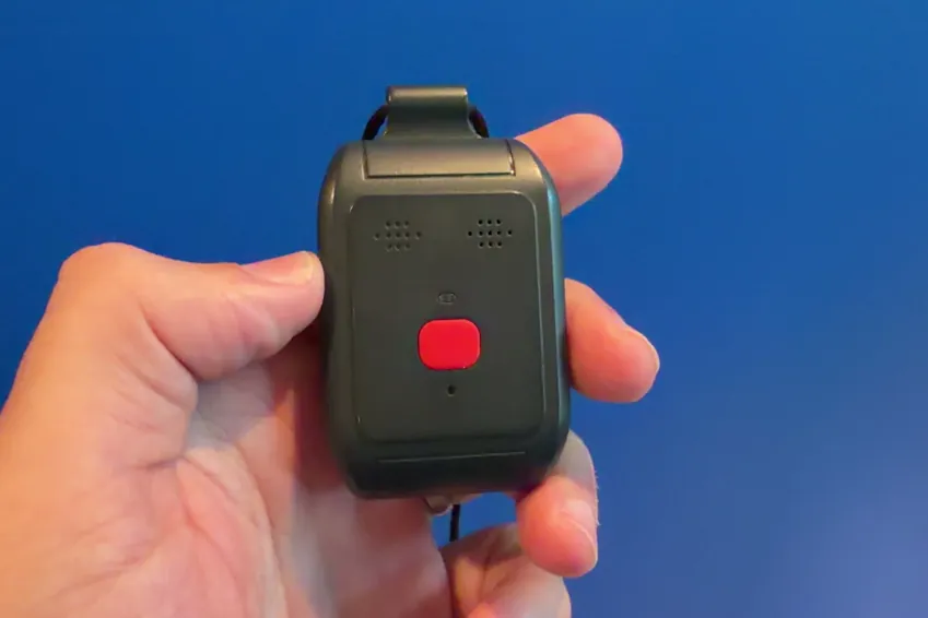 Alert1 LifeBeacon pendant held in a hand against a blue background, showing the red help button and speaker grilles