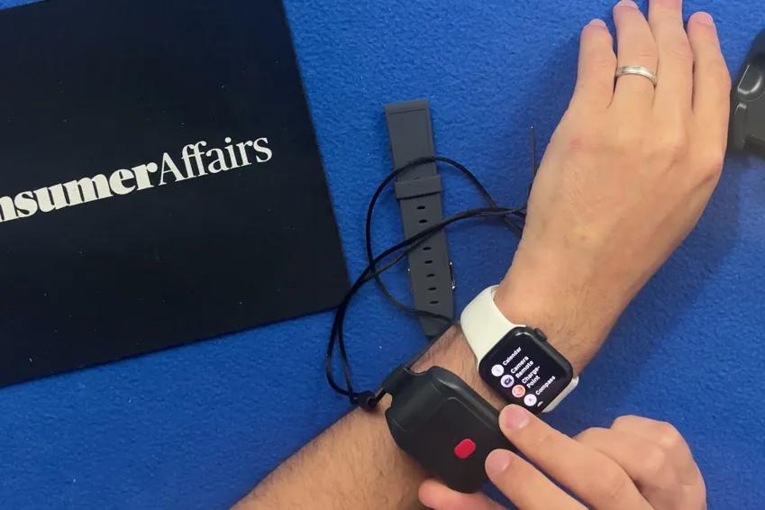 Alert1 LifeBeacon pendant held on a wrist next to an Apple Watch for size, beside a ConsumerAffairs mousepad