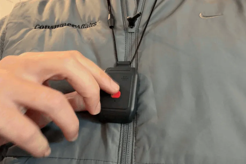 A finger pressing and holding the red help button on the LifeBeacon pendant worn on the chest
