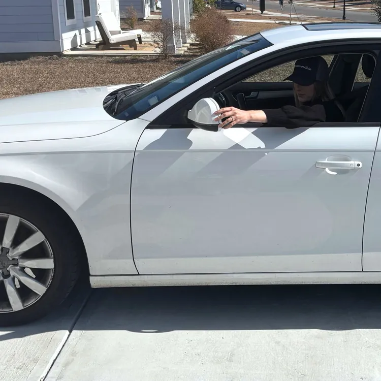 White sedan parked on a driveway with a person inside holding a phone.