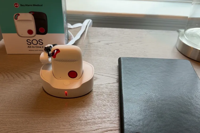 SOS All-in-One 2 pendant sitting in its charging cradle on a bedside table, the Bay Alarm Medical box behind it