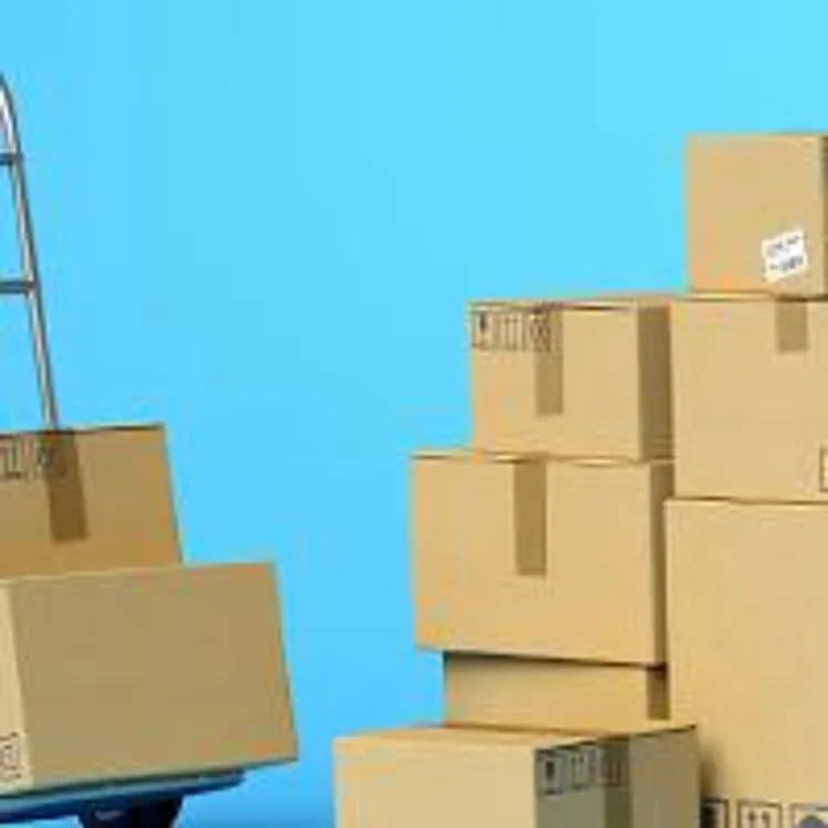 two cardboard boxes on hand truck beside a stack of other cardboard boxes