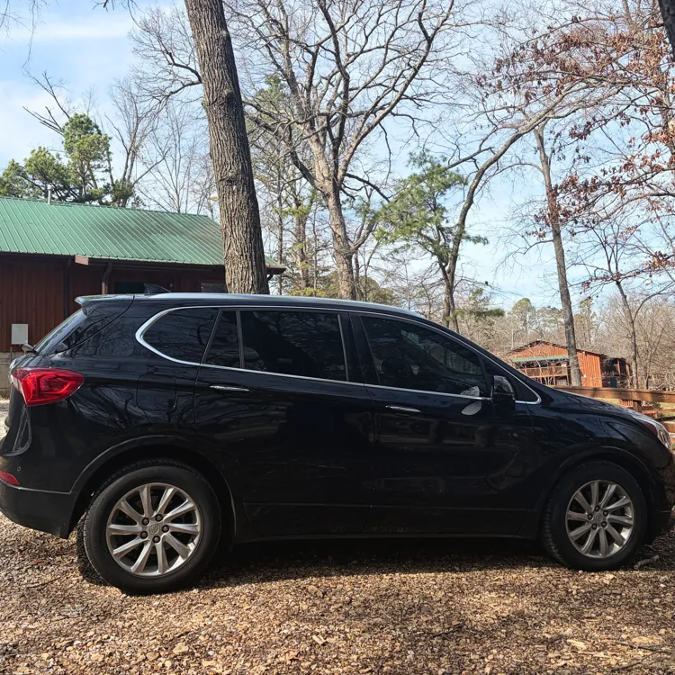 Black SUV parked on a dirt path surrounded by bare trees and a wooden cabin with a green roof in the background.