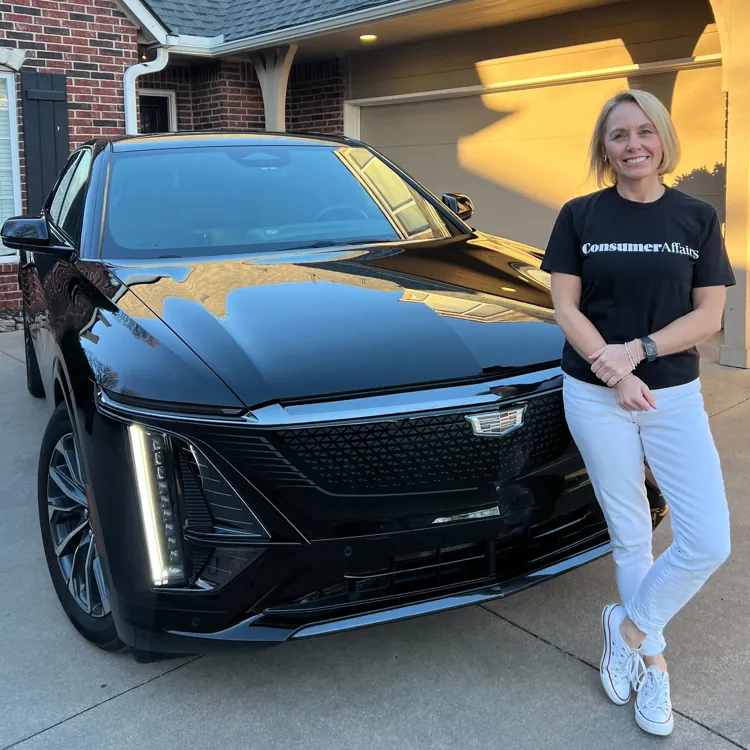 Person standing next to a black car parked in a driveway of a suburban home with brick and siding exterior.