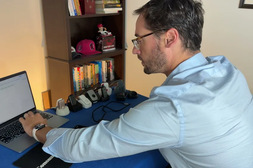 Chris Kolmar at a laptop testing medical alert devices, with several pendants and charging docks lined up on the table