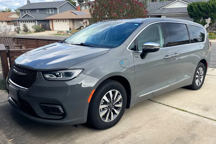 Gray minivan parked in a suburban driveway with houses and trees in the background.