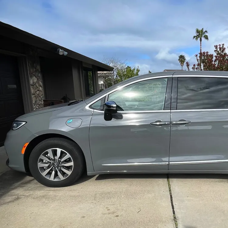 Gray minivan parked in a suburban driveway under a cloudy sky with trees and houses in the background.