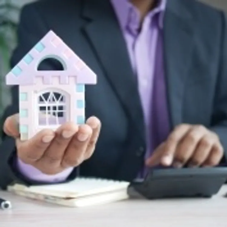 Man in suit holding a small house model at a desk with a calculator