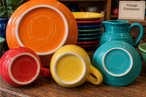 Assorted colorful vintage Fiesta ceramic mugs, plates, and a pitcher displayed on a wooden shelf