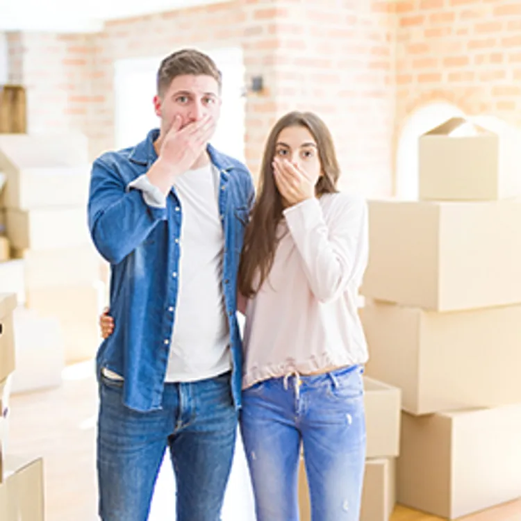 newly moved couple with hand covering their mouth with boxes in the background