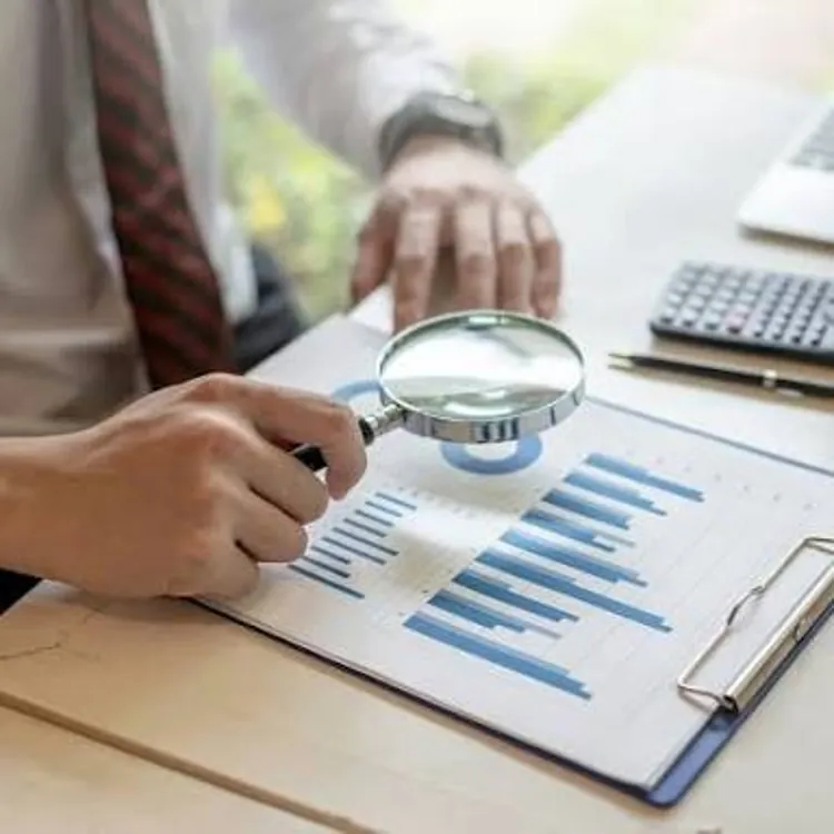 Person examining a financial graph on a clipboard with a magnifying glass at a desk