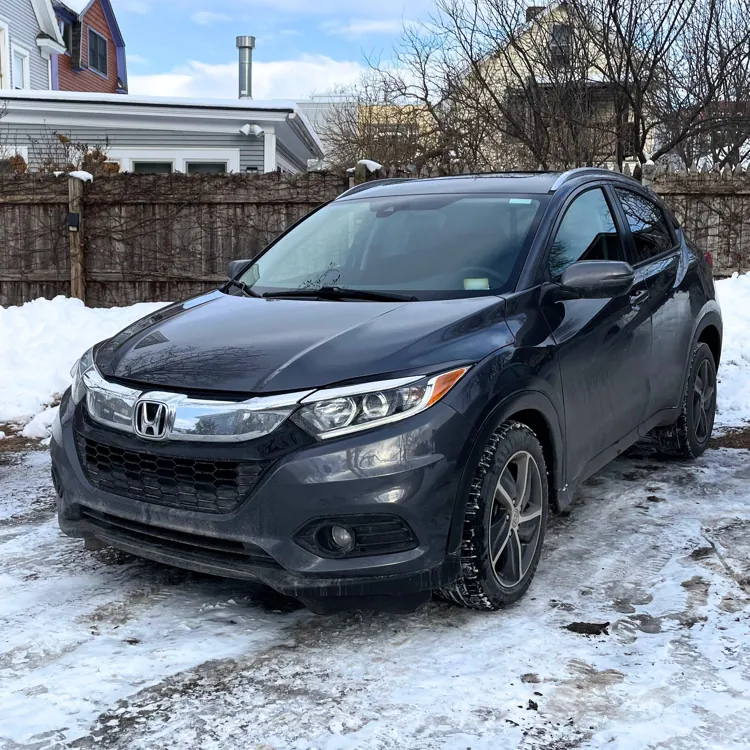 Black SUV parked on a snowy driveway in front of a wooden fence and residential houses.