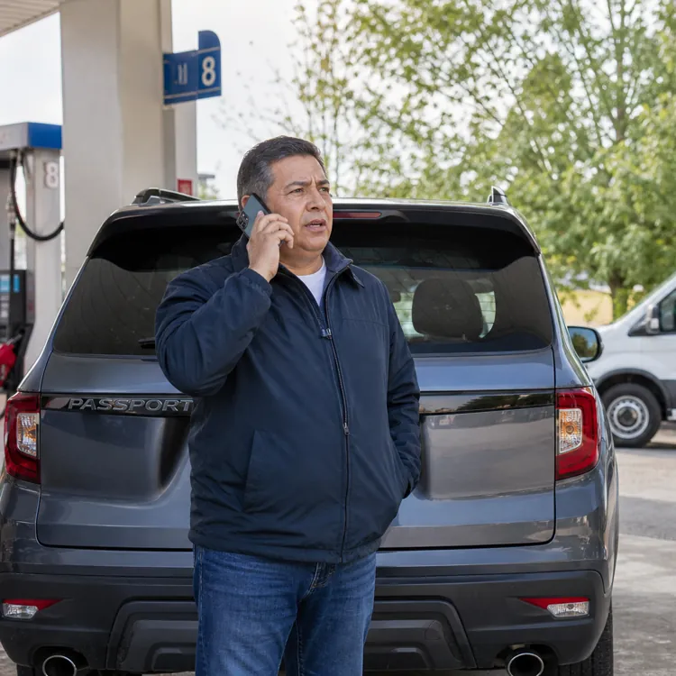 Man on phone standing by SUV at gas station with Honda roadside assistance van in background