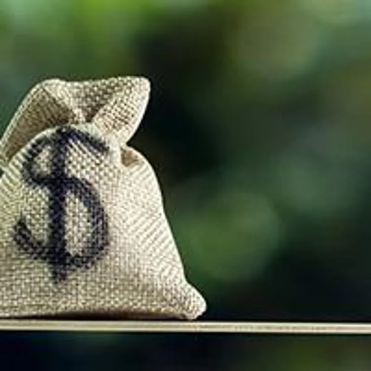 us dollar in sack bag and wooden house model on opposite ends of a scale with green tree bokeh as background
