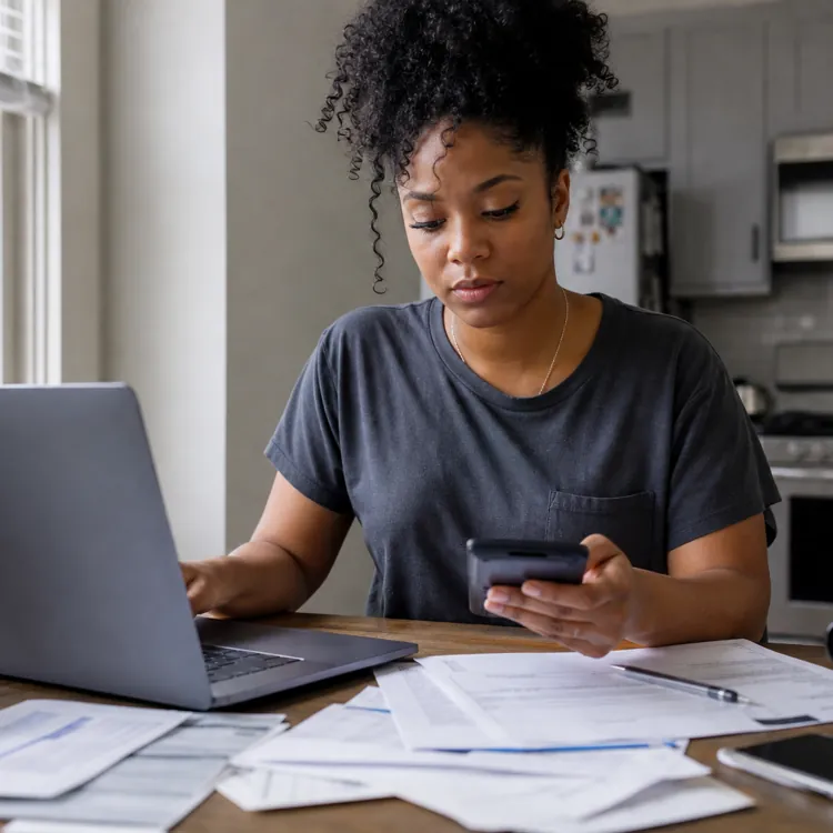 Woman using laptop and phone at kitchen table with bills and paperwork