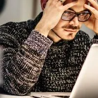 a young man wearing glasses is sitting in front of his laptop while holding his head and pondering something