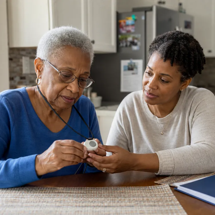 Older woman and caregiver examining a medical alert device at a kitchen table