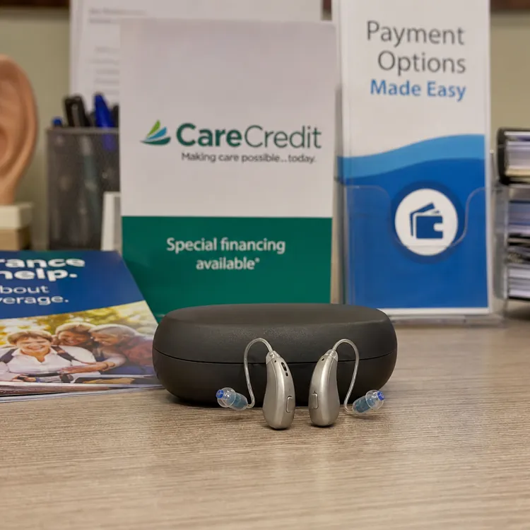 Two hearing aids and a case on a desk with brochures and payment option signs in the background