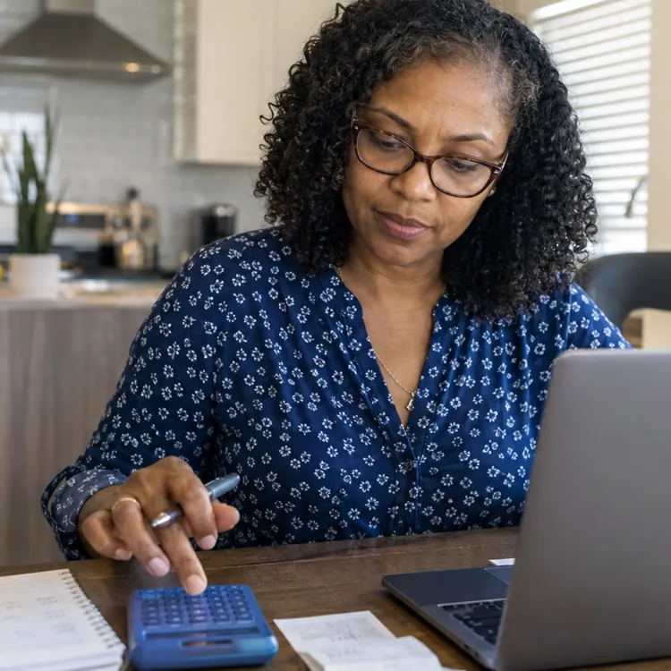 Woman using calculator and laptop at a kitchen table with papers and coffee mug