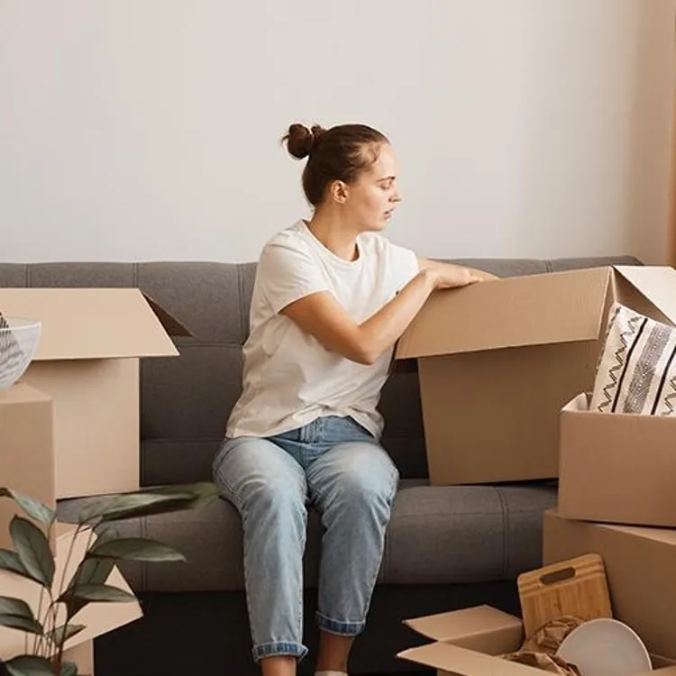 A woman unpacks cardboard boxes in a living room with a sofa and household items.