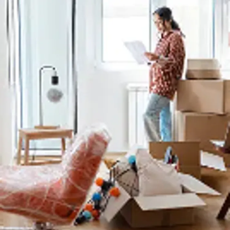 Woman standing in front of a window in a house, next to stacked boxes, opened box with pillows, and a side table with a lamp.