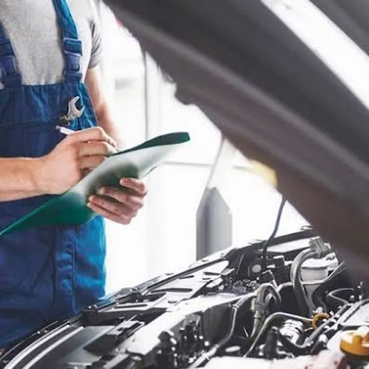 Mechanic in blue overalls holding a clipboard inspecting a car engine with the hood open