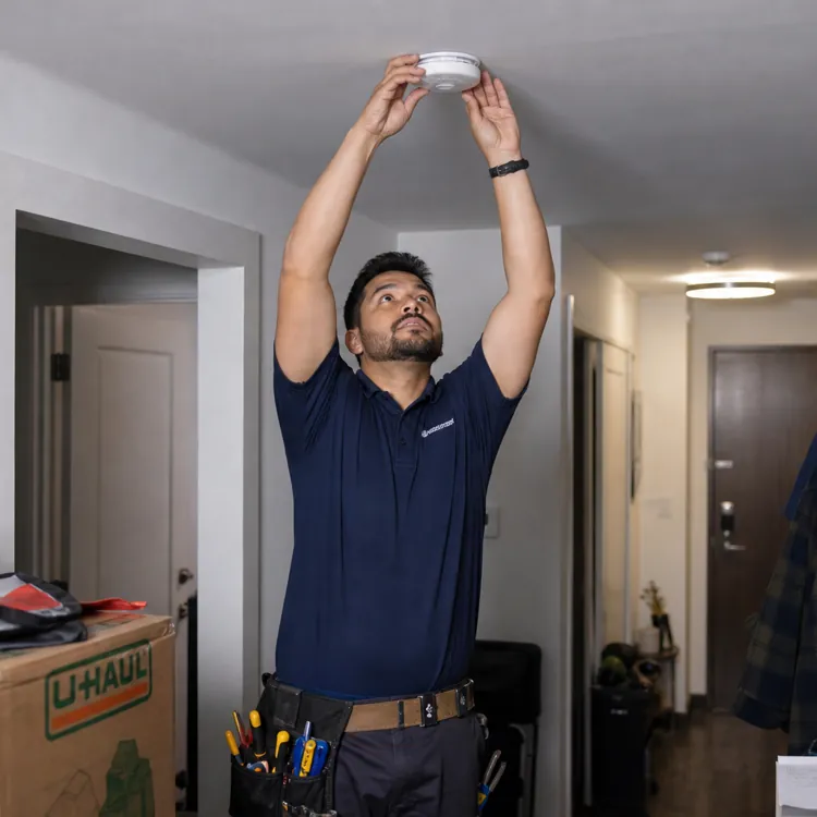 Maintenance worker installing a smoke detector on a hallway ceiling near moving boxes