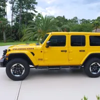 Yellow Jeep Rubicon parked on a driveway surrounded by palm trees and greenery under a partly cloudy sky.