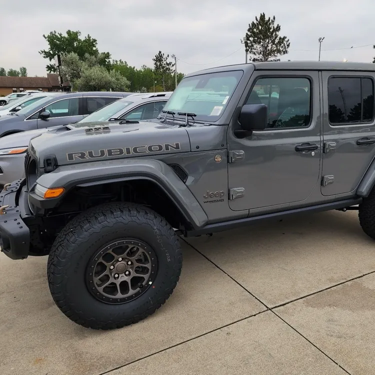 Gray Jeep Rubicon parked in a lot with other vehicles, surrounded by trees and buildings.