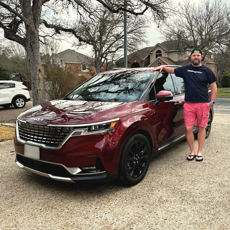 Person standing next to a red car parked in a suburban driveway with trees and houses in the background.