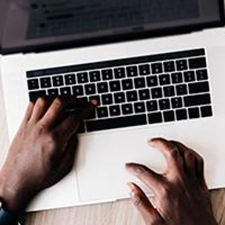 pair of hands typing on a laptop with pen and paper on the table on the side