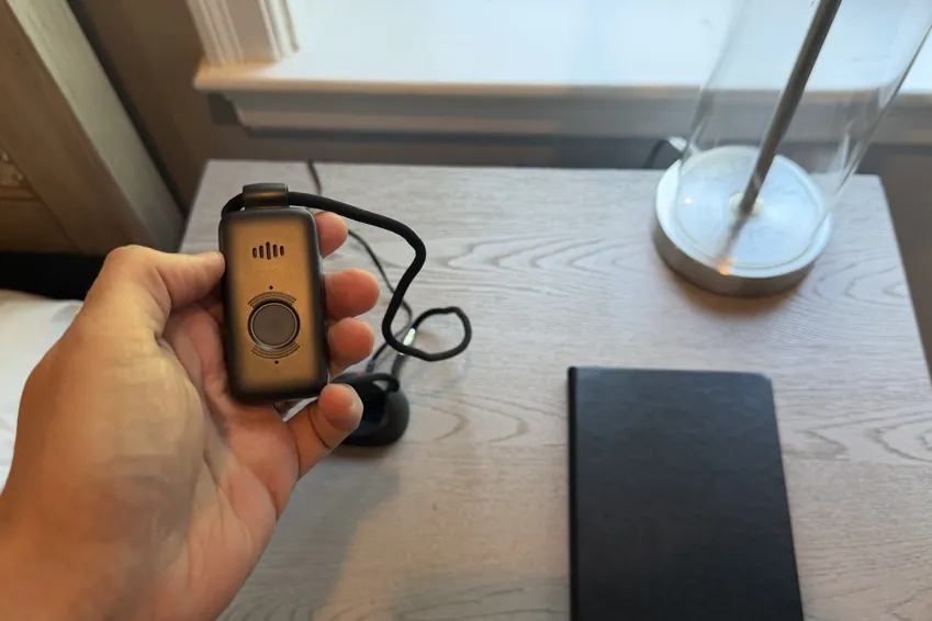 LifeFone VIP X2 pendant held in a hand above a bedside table, its charging dock in the background