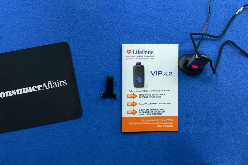 Overhead view of the LifeFone VIP X2 pendant on its lanyard in the charging dock, belt clip and user guide beside a ConsumerAffairs mousepad