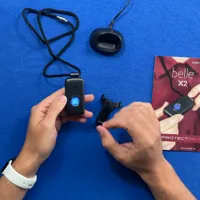 Hands holding a medical alert device with accessories, user guide, and charging dock on a blue surface