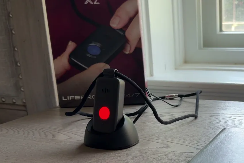 Belle X2 pendant in its charging cradle on a bedside table with a red light on, the box behind it