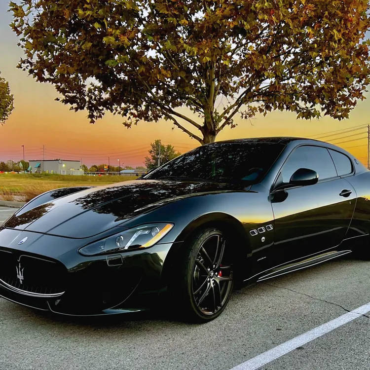 Black sports car parked in a lot with a tree in the background and a colorful sunset sky.