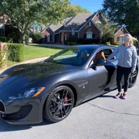 Person standing next to a black luxury sports car parked on a suburban street with brick houses and green lawns.