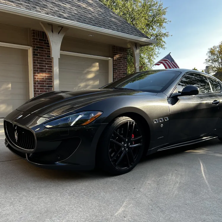 A sleek black luxury sports car parked on a driveway with an American flag in the background on a sunny day.