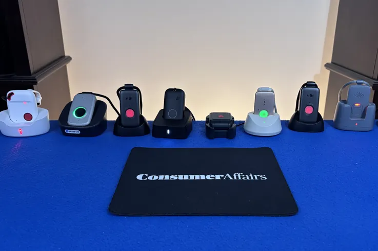 A lineup of medical alert pendants sitting on their charging bases along a blue table behind a ConsumerAffairs mousepad