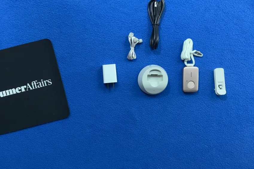 Overhead view of the MGMini pendant on its lanyard, charging base, USB-A wall adapter, charging cable, spare adjustable lanyard and belt clip beside a ConsumerAffairs mousepad
