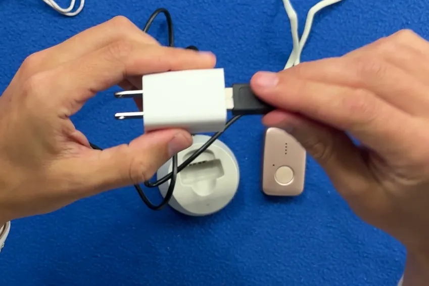 Hands plugging the MGMini charging cable into its USB-A wall adapter above the charging base and pendant