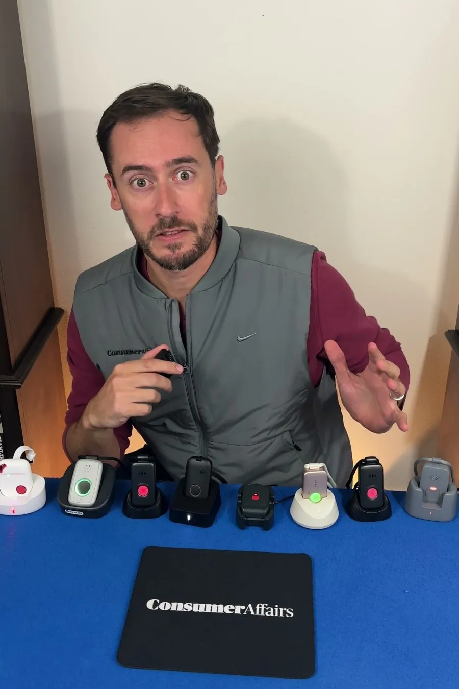Chris Kolmar at a blue table with nine medical alert pendants on their chargers and a ConsumerAffairs mouse pad