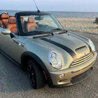 A silver Mini Cooper convertible with black racing stripes parked near a beach, featuring tan leather seats.
