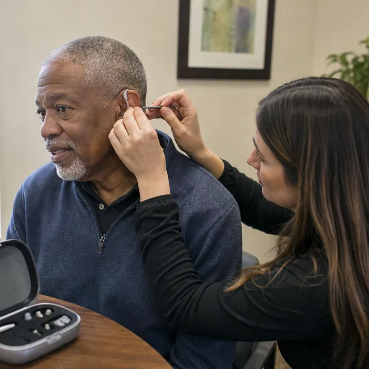 Audiologist fitting a hearing aid on an older man in a clinic with a hearing aid case on the table