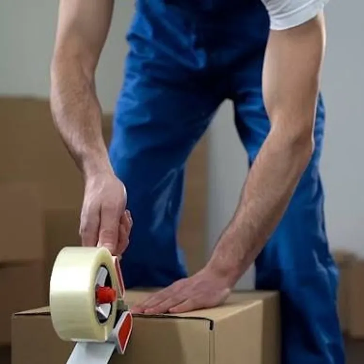 Person sealing a cardboard box with packing tape in a room with several boxes