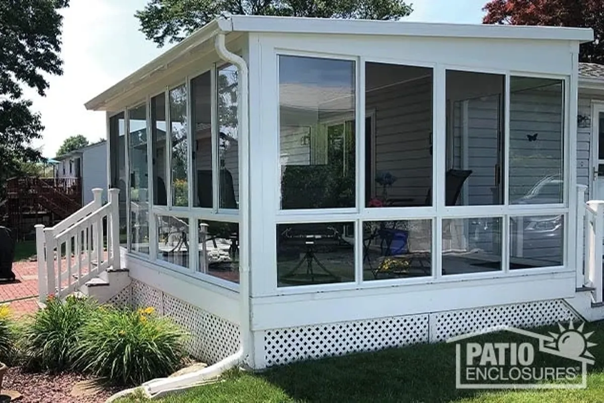 A white-framed Patio Enclosures sunroom addition on the back of a suburban home, with glass walls on three sides looking onto a landscaped garden.