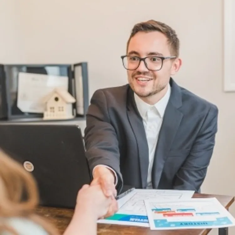 Man in suit shaking hands across desk with papers and laptop