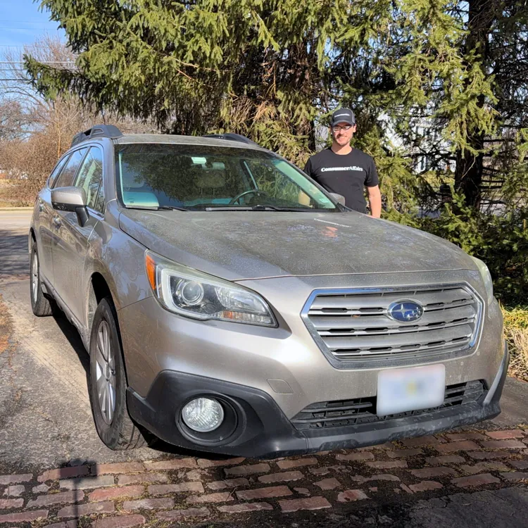 A person standing next to a silver Subaru car parked on a driveway with trees in the background on a sunny day.