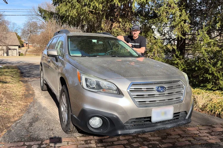 Person leaning on a silver car parked on a driveway with trees and houses in the background on a sunny day.