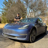 Person standing next to a blue electric car parked on a driveway with trees in the background on a sunny day.